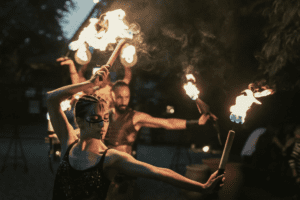 Performer con trucco tribale ed espressione concentrata durante uno spettacolo di fuoco serale, con torce infuocate mosse in sincronia davanti a un pubblico immerso nell'atmosfera suggestiva di The Sanctuary